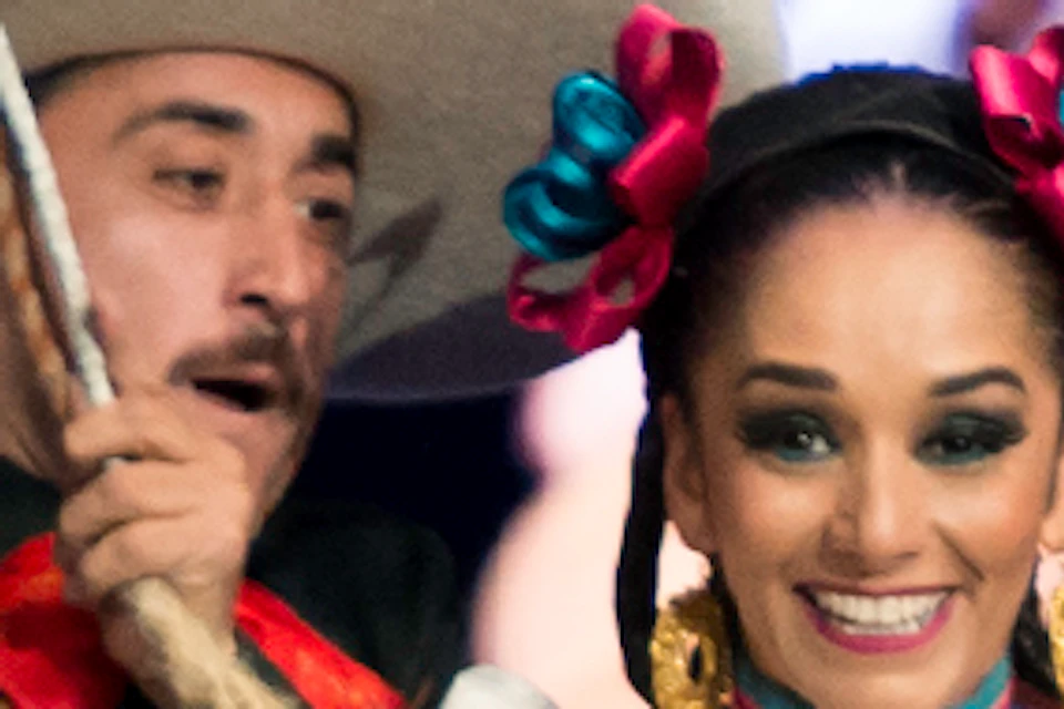 A man in traditional charro attire spins a lasso around a woman in a colorful folkloric dress, with a mariachi band playing in the background.