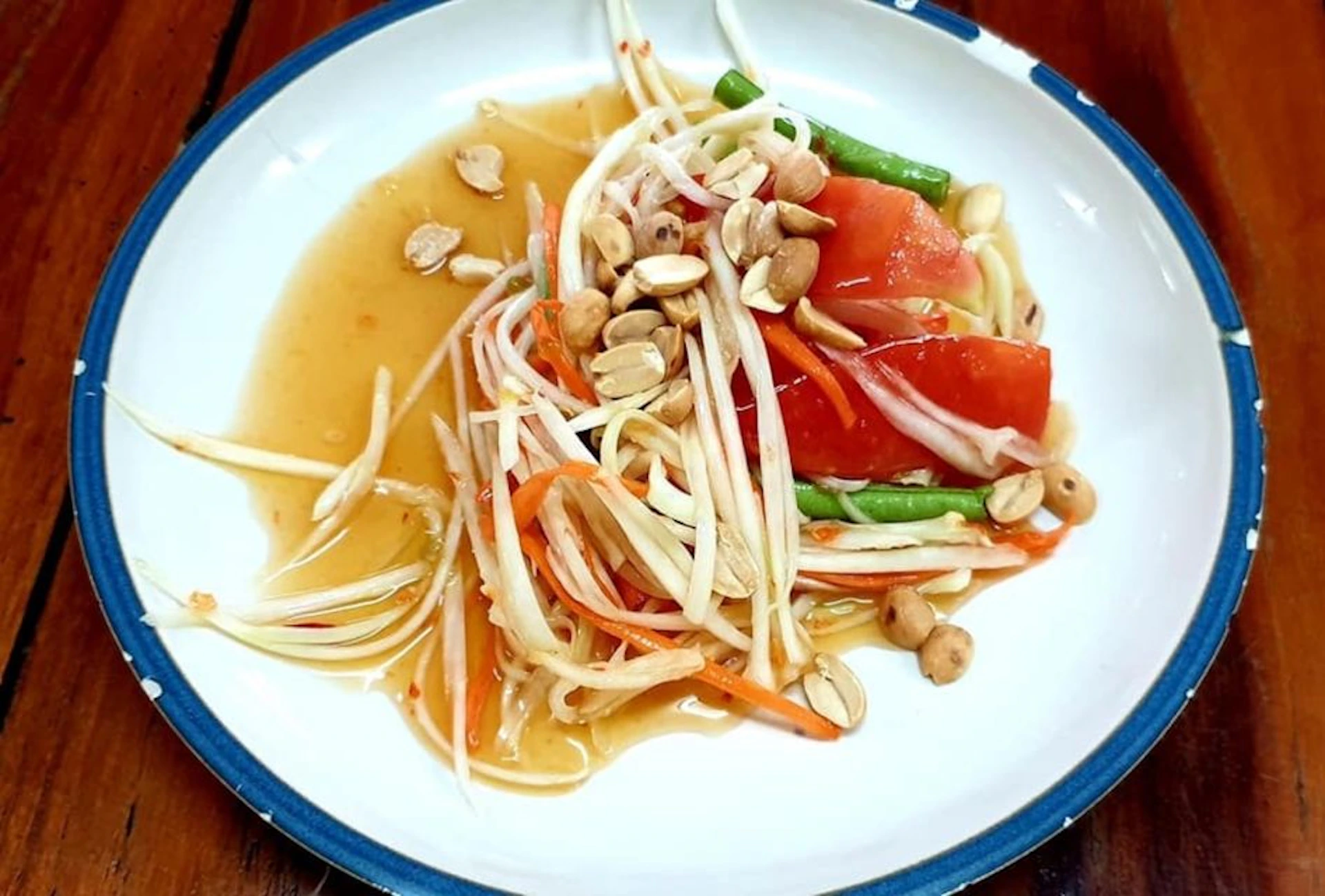 A plate of Thai papaya salad with shredded papaya, tomatoes, green beans, peanuts, and a light dressing.