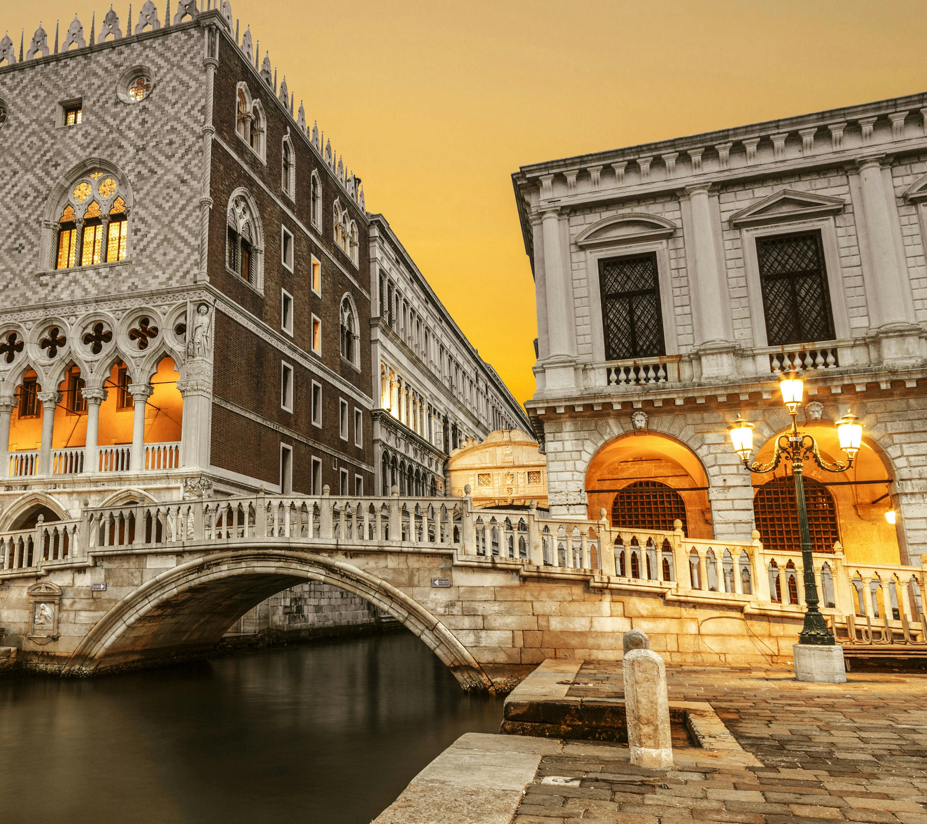 Zdobený most a historické budovy podél kanálu při západu slunce s osvětlenými pouličními lampami a teplými žlutými okny.