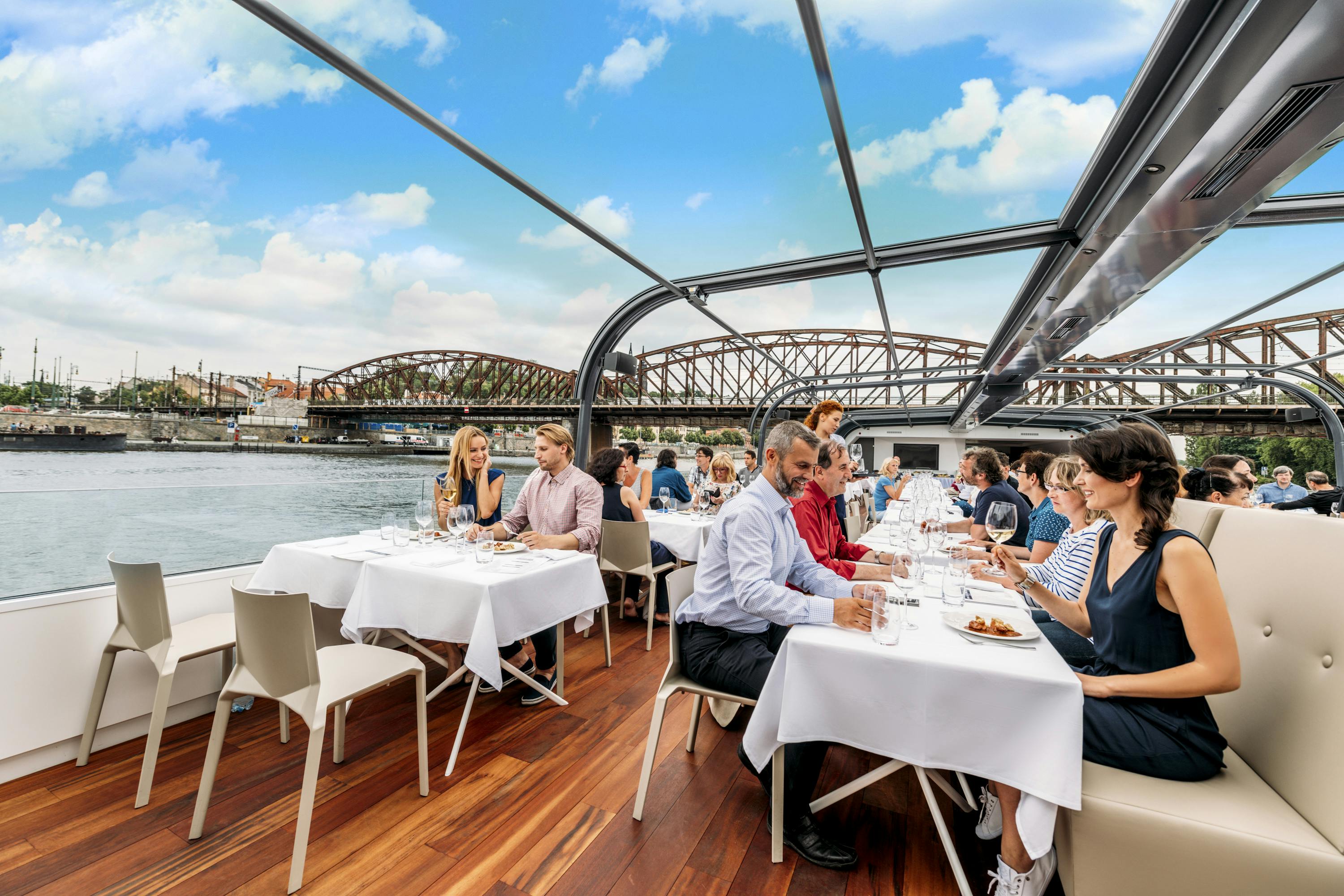 People dining on a boat with white tablecloths, wooden floor, and a glass roof. A bridge spans the river in the background.