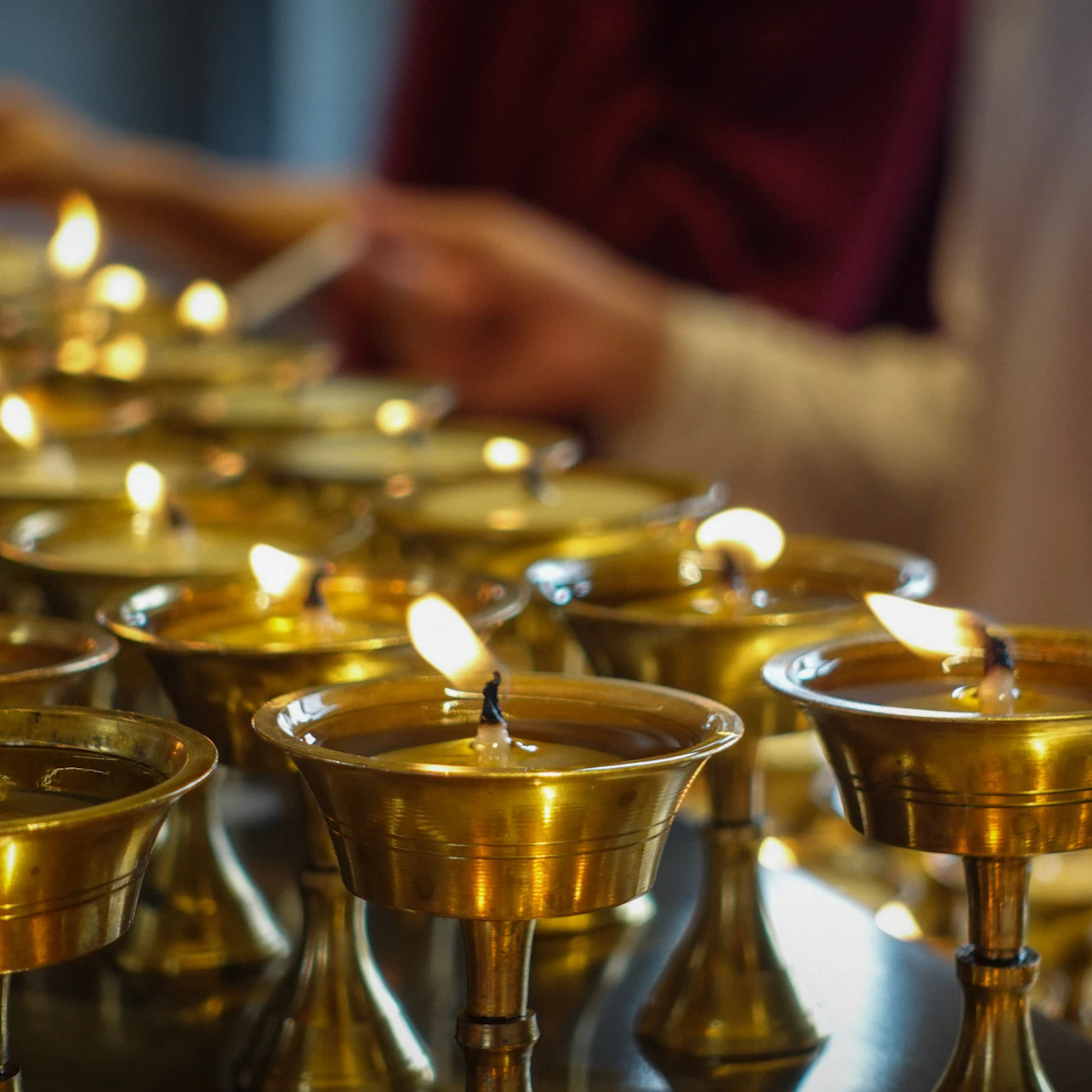 Brass oil lamps with small flames arranged in rows, with blurred figures in the background.