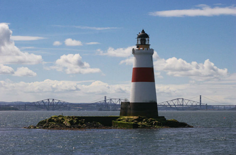 White lighthouse with a red stripe on a small rocky island, surrounded by water under a partly cloudy sky with a distant bridge.