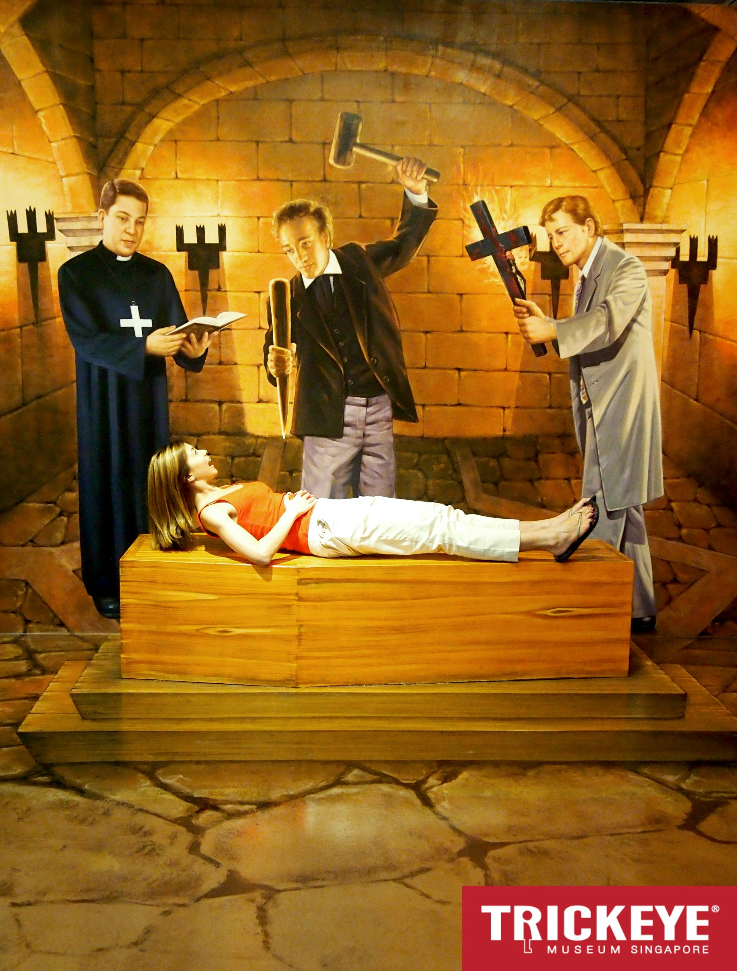 A woman lies on a wooden box while three men, one in a robe holding a book, another with a mallet and stake, stand nearby in a stone room.