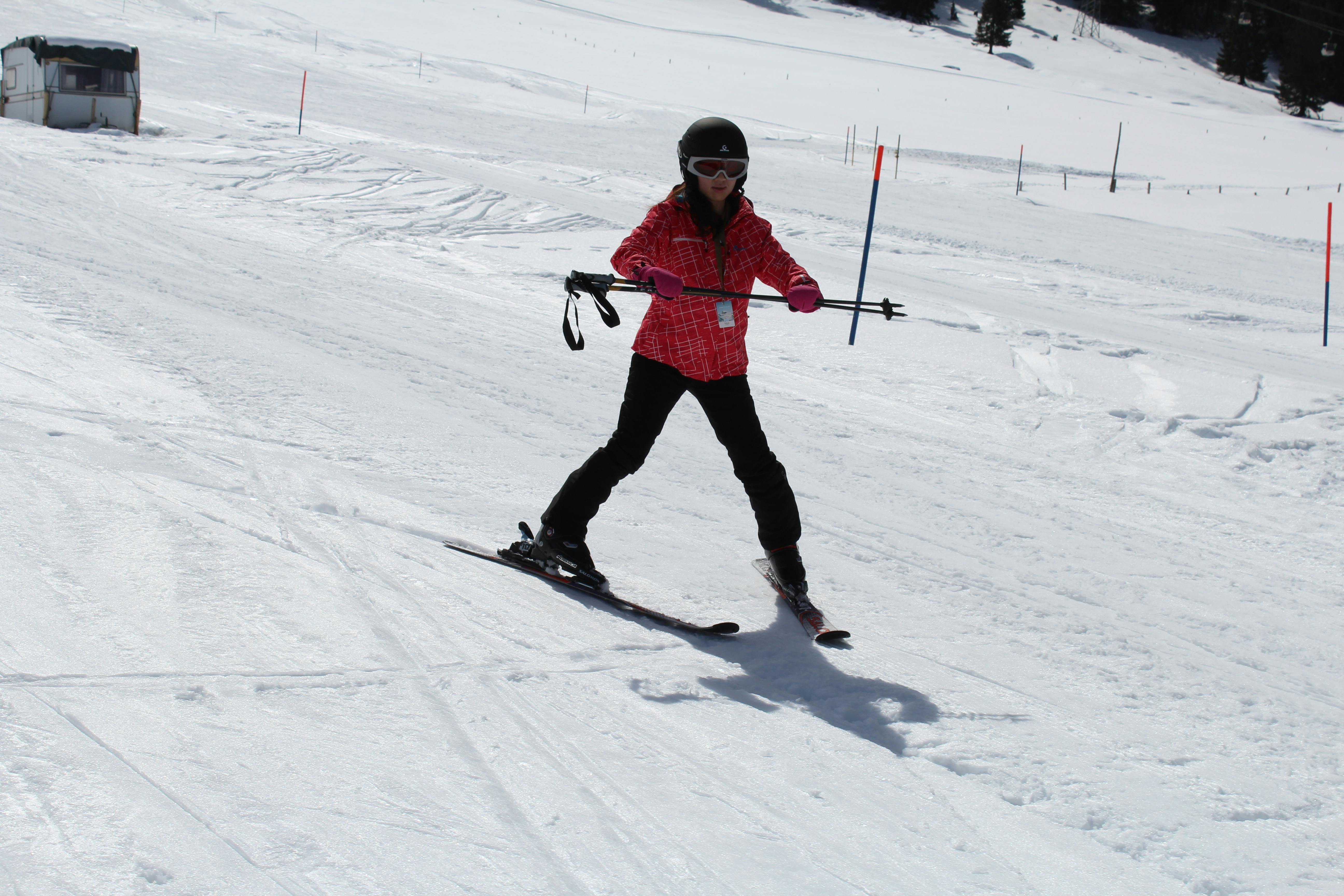 Άτομο που κάνει σκι σε χιονισμένη πλαγιά, φορώντας κόκκινο μπουφάν, μαύρο παντελόνι, κράνος και γυαλιά, κρατώντας μπαστούνια του σκι.