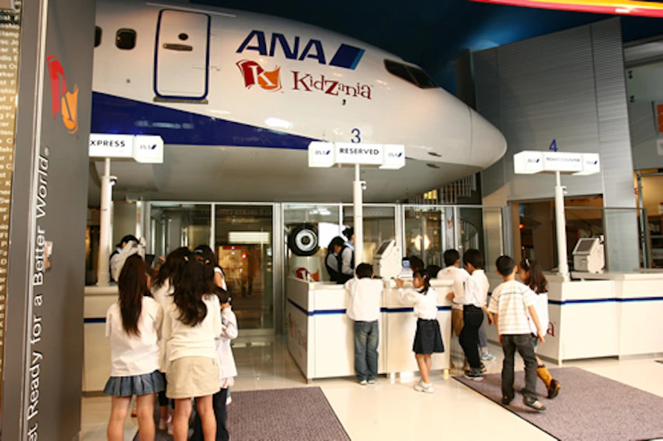 Children in line at a simulated airport check-in area with an ANA airplane above, inside KidZania.