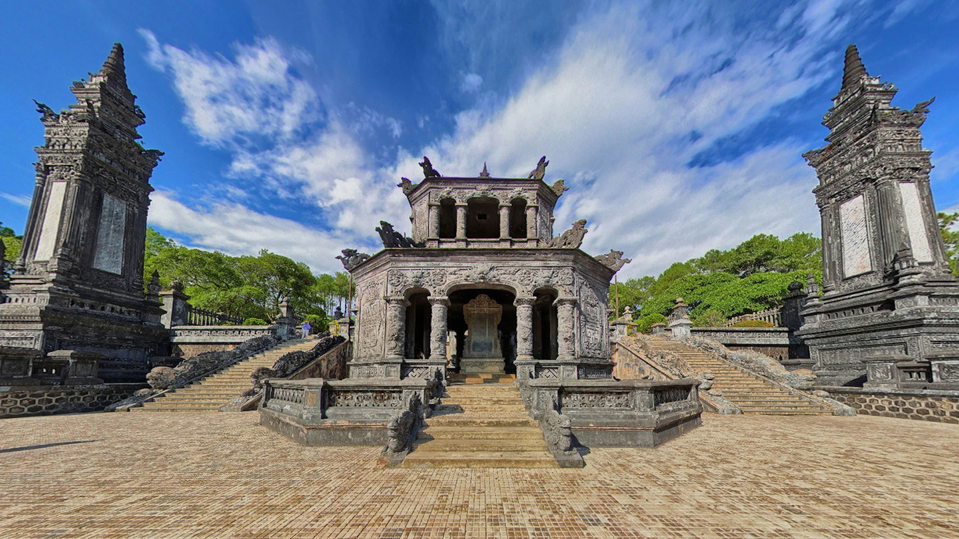 An ornate stone temple with intricate carvings, surrounded by stairs and greenery under a partly cloudy blue sky.