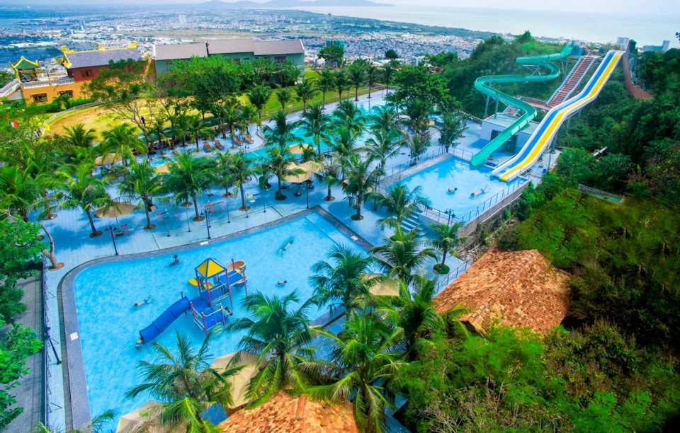 A water park with pools, tall water slides, palm trees, and surrounding greenery, viewed from an elevated perspective.