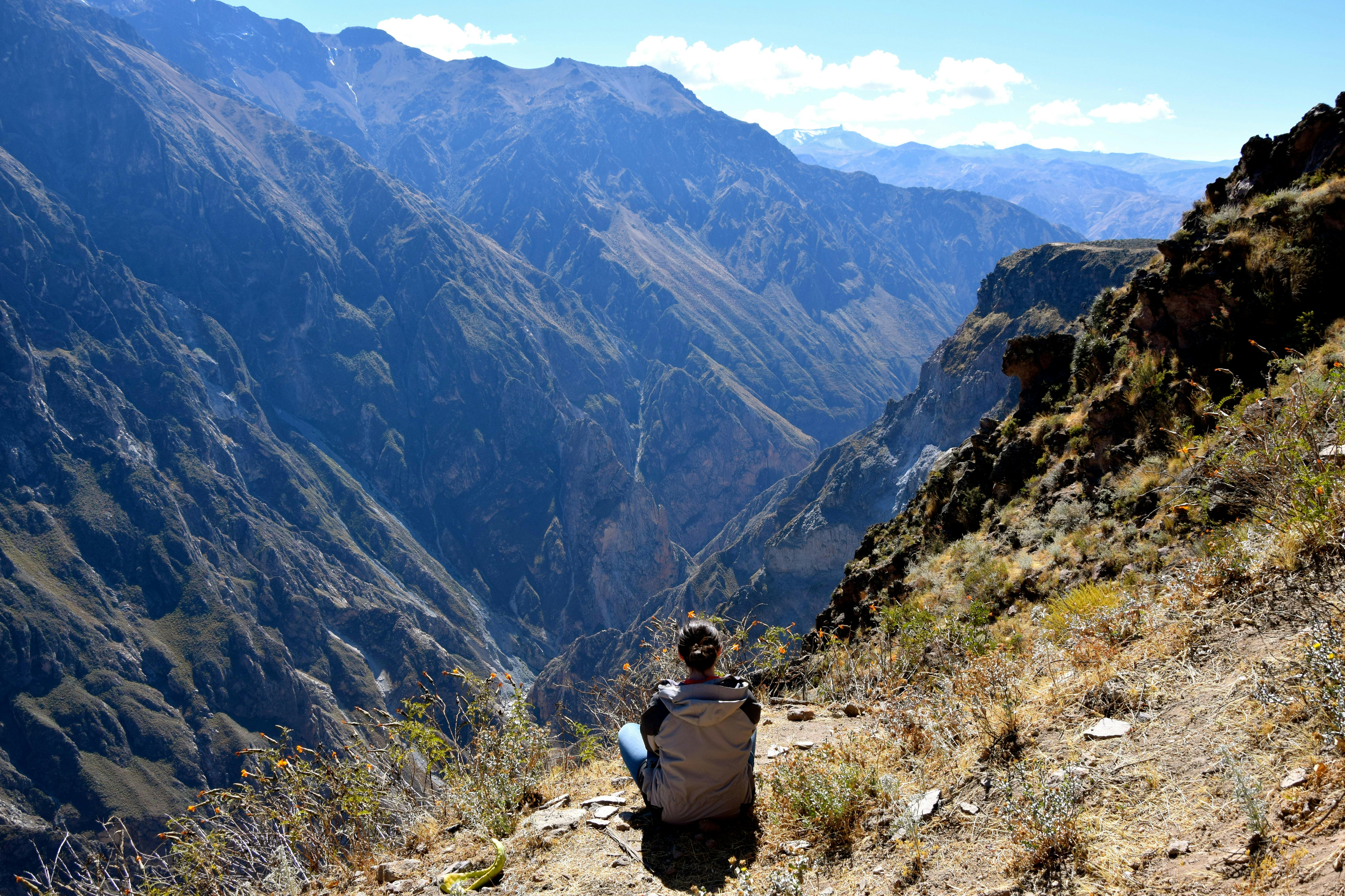 Человек, сидящий на краю скалы, с видом на обширный горный пейзаж под ясным солнечным небом.