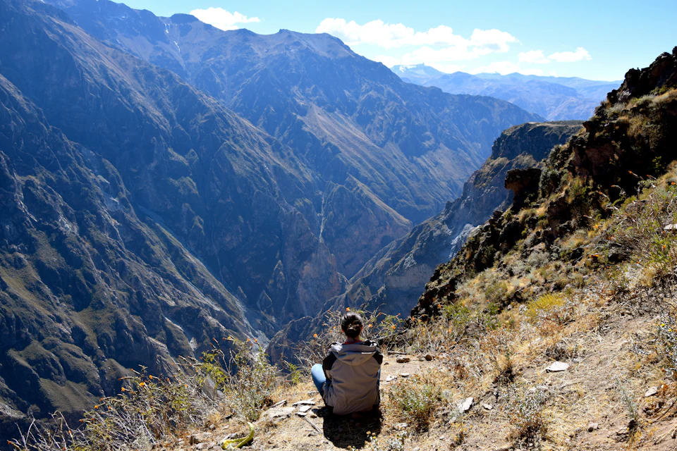 Person sitting on the edge of a cliff, overlooking a vast mountainous landscape under a clear, sunny sky.