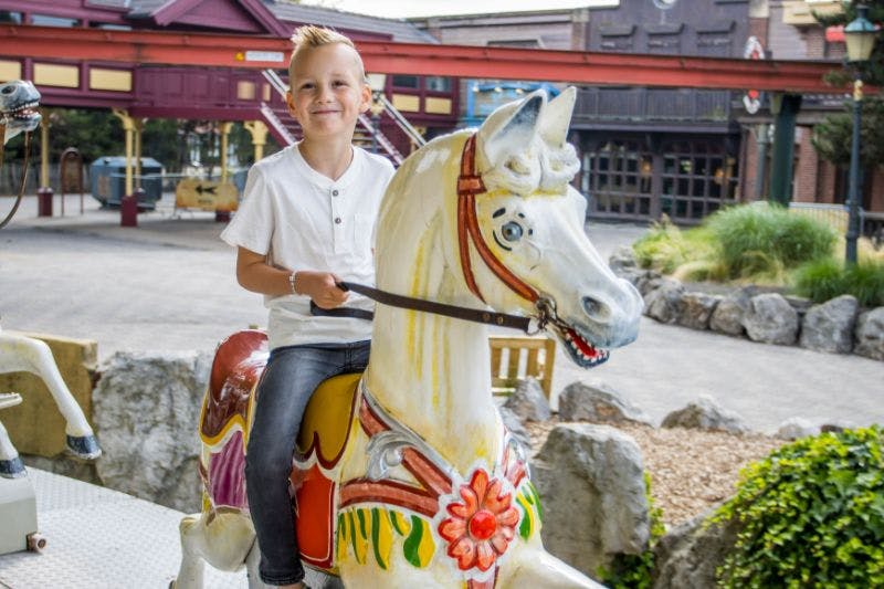 A boy smiling while riding a painted carousel horse, with a background of a building and outdoor fairground.