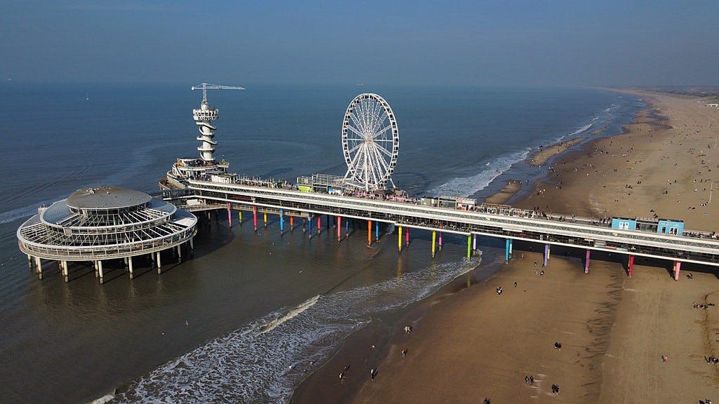 Molo della spiaggia con ruota panoramica, torre di osservazione e struttura circolare sull'acqua. I frequentatori della spiaggia passeggiano lungo l'arenile.