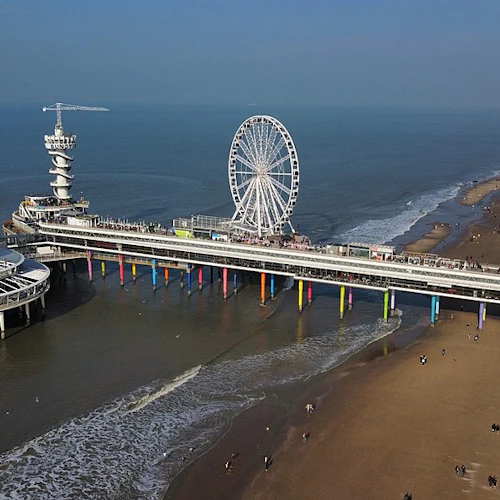 Strandpir med pariserhjul, observationstorn och cirkulär struktur över vattnet. Strandgäster promenerar längs sandstranden.