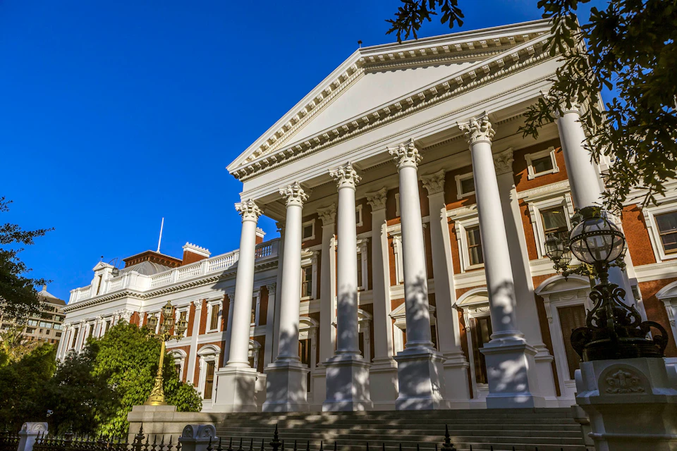 A neoclassical building with tall white columns, a pediment, and windows, surrounded by trees under a clear blue sky.
