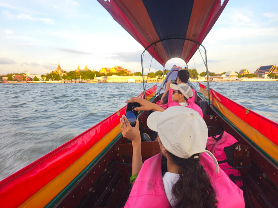 People in pink life jackets and white caps seated in a boat on a river, taking photos of distant golden temple structures.
