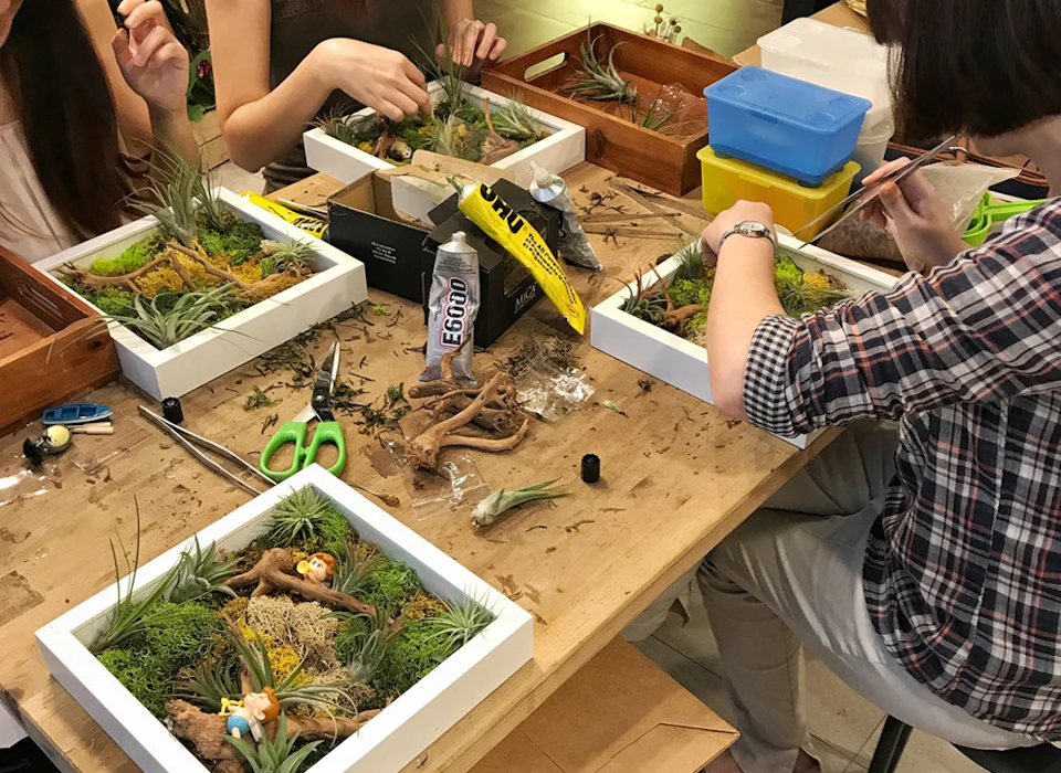 People sitting around a table crafting terrariums with plants, moss, and decorations in shallow wooden boxes.