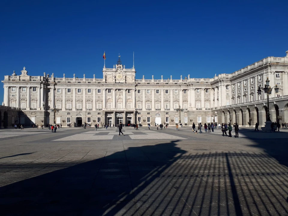 A large, ornate white building with multiple windows and a clock tower stands under a clear blue sky. People are walking in the foreground.