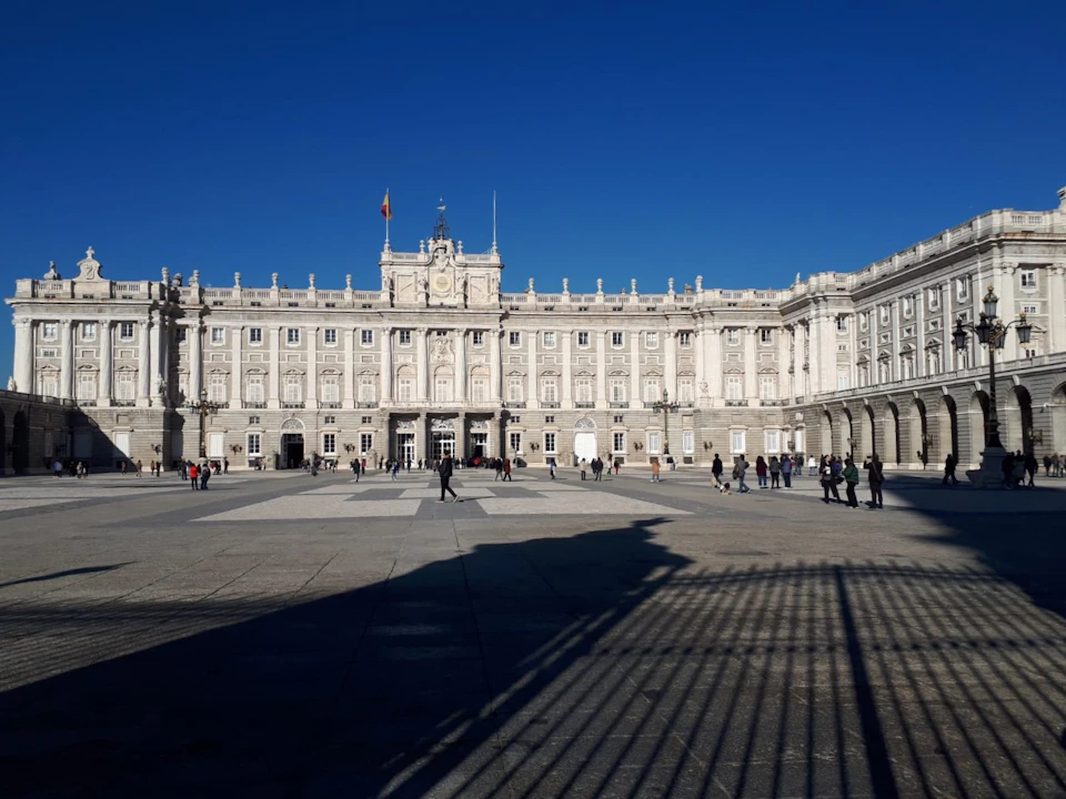 A large, ornate white building with multiple windows and a clock tower stands under a clear blue sky. People are walking in the foreground.