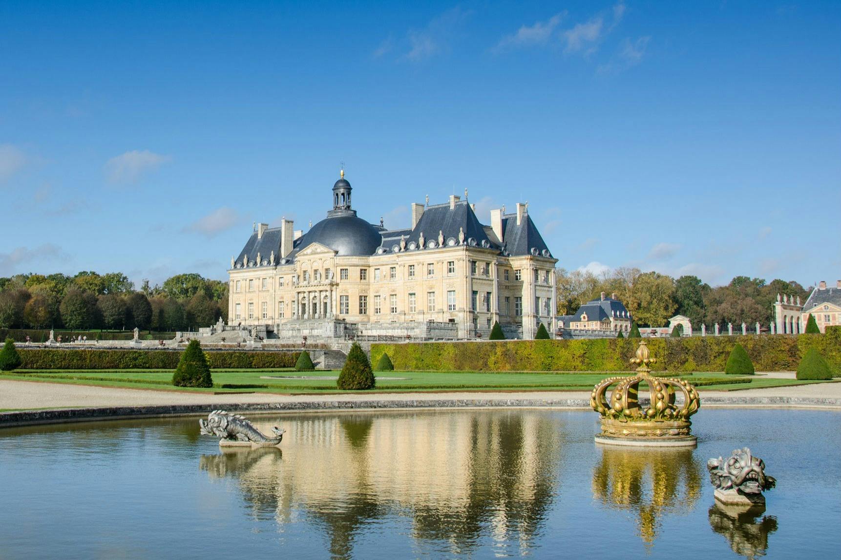 A grand palace with blue-domed roofs reflected in a large ornamental pond, featuring sculptures and a crown-shaped fountain.