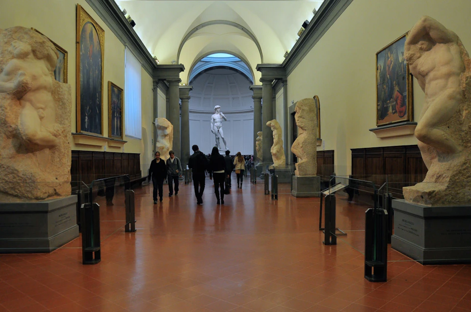 People walking through an art gallery corridor with large sculptures on both sides and the statue of David at the end.