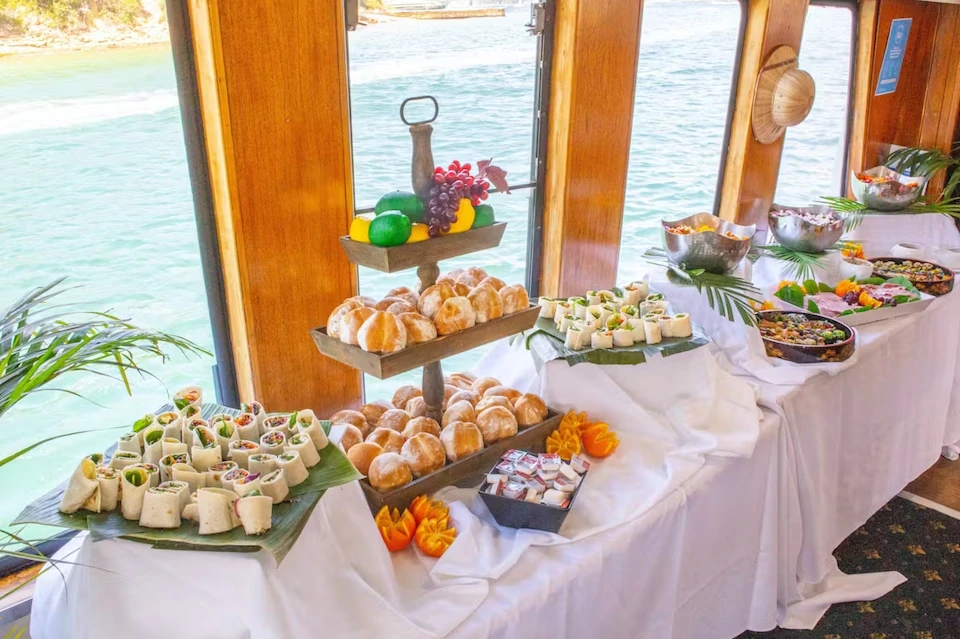 Buffet table on a boat with assorted appetizers including wraps, bread rolls, fruit, and dips, against a background of large windows showing water outside.