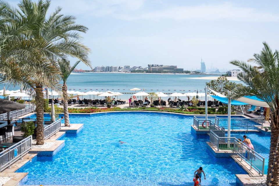 A pool with swimmers, palm trees, lounge chairs, umbrellas, and a view of the ocean and city buildings in the background.