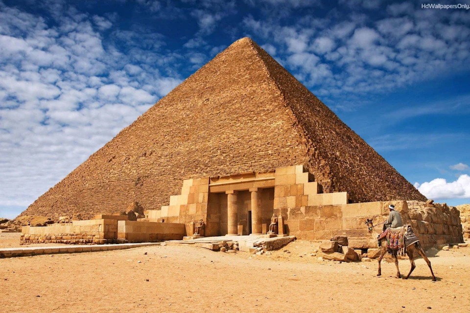 The Great Pyramid of Giza under a partly cloudy sky, with the entrance and surrounding sandy landscape visible.