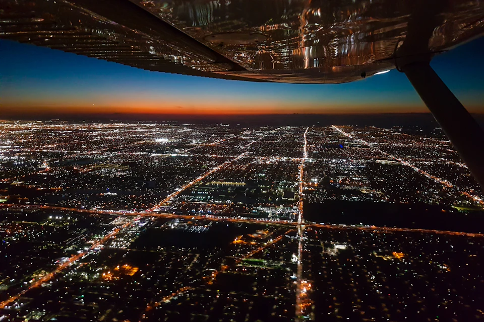 Aerial view of a city at dusk, with illuminated streets and buildings, seen from an airplane window.