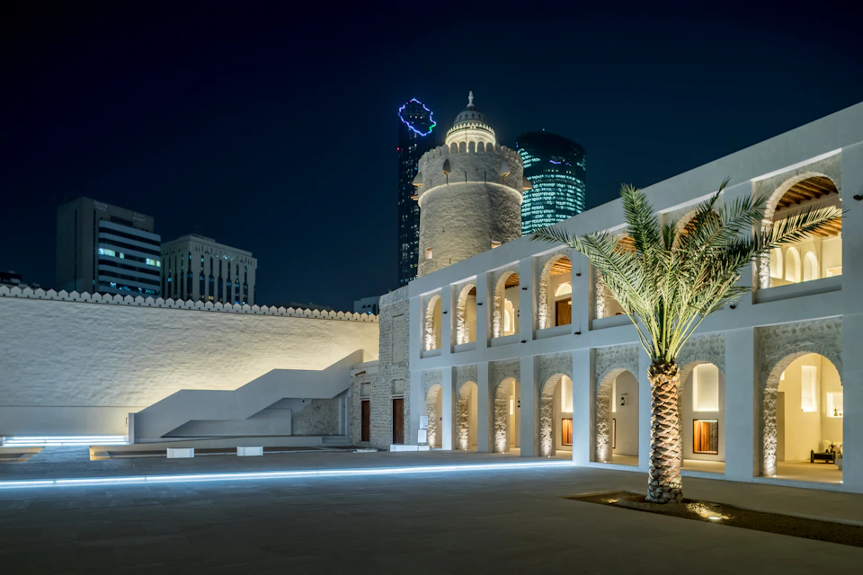 Stone building with arched doorways and a central dome lit at night, palm tree in the foreground, modern buildings in the background.