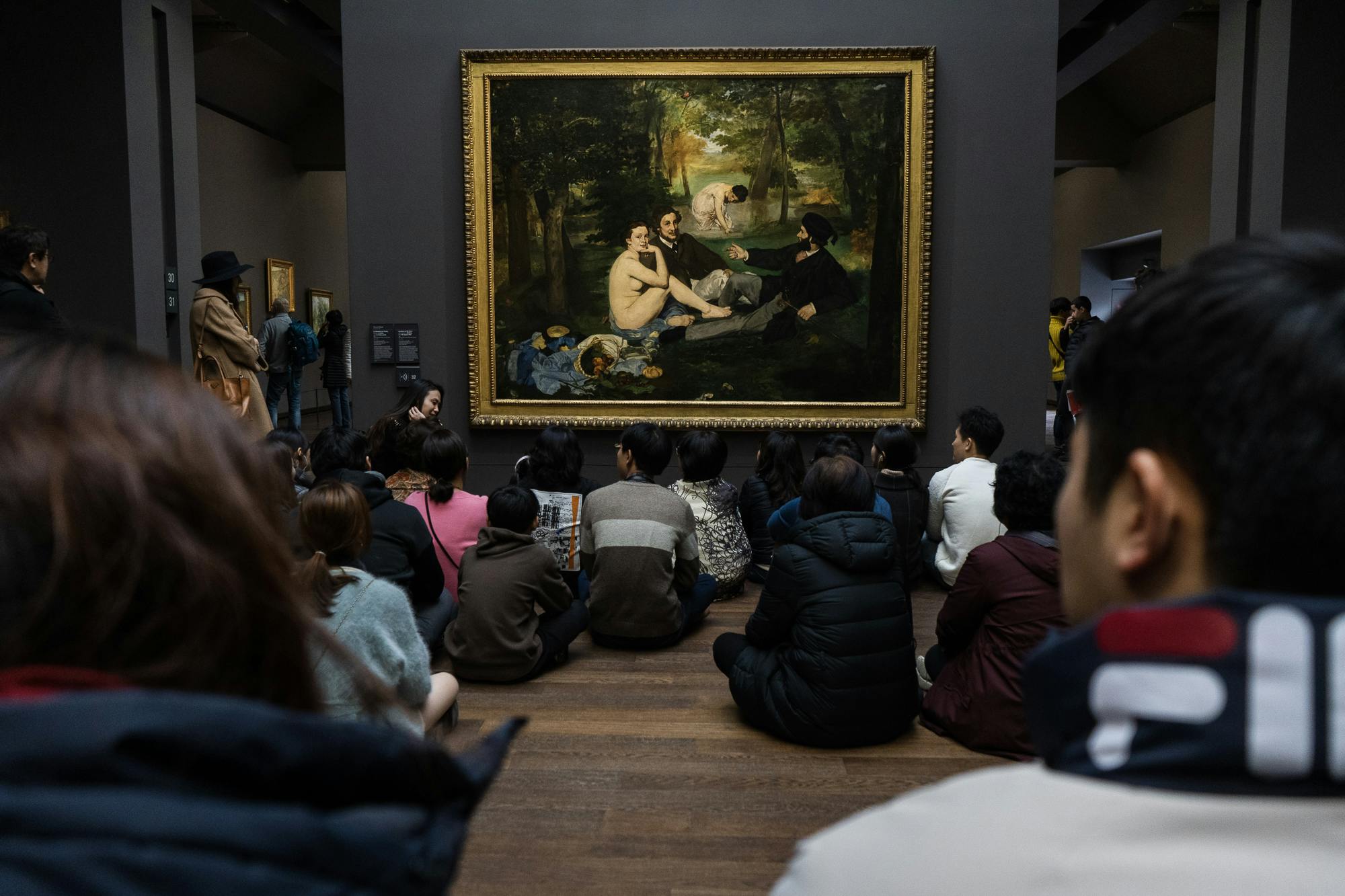 A group of people sit on the floor, viewing a large painting of people in a forest, displayed in a museum.