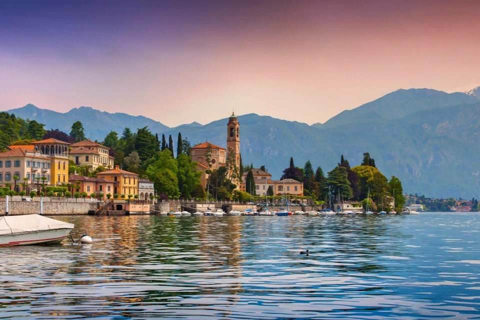 A picturesque lakeside town with colorful buildings, a bell tower, trees, and mountains in the background at sunset.