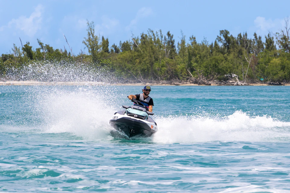 A person riding a jet ski on a blue body of water near a wooded shoreline, creating splashes as they speed along.