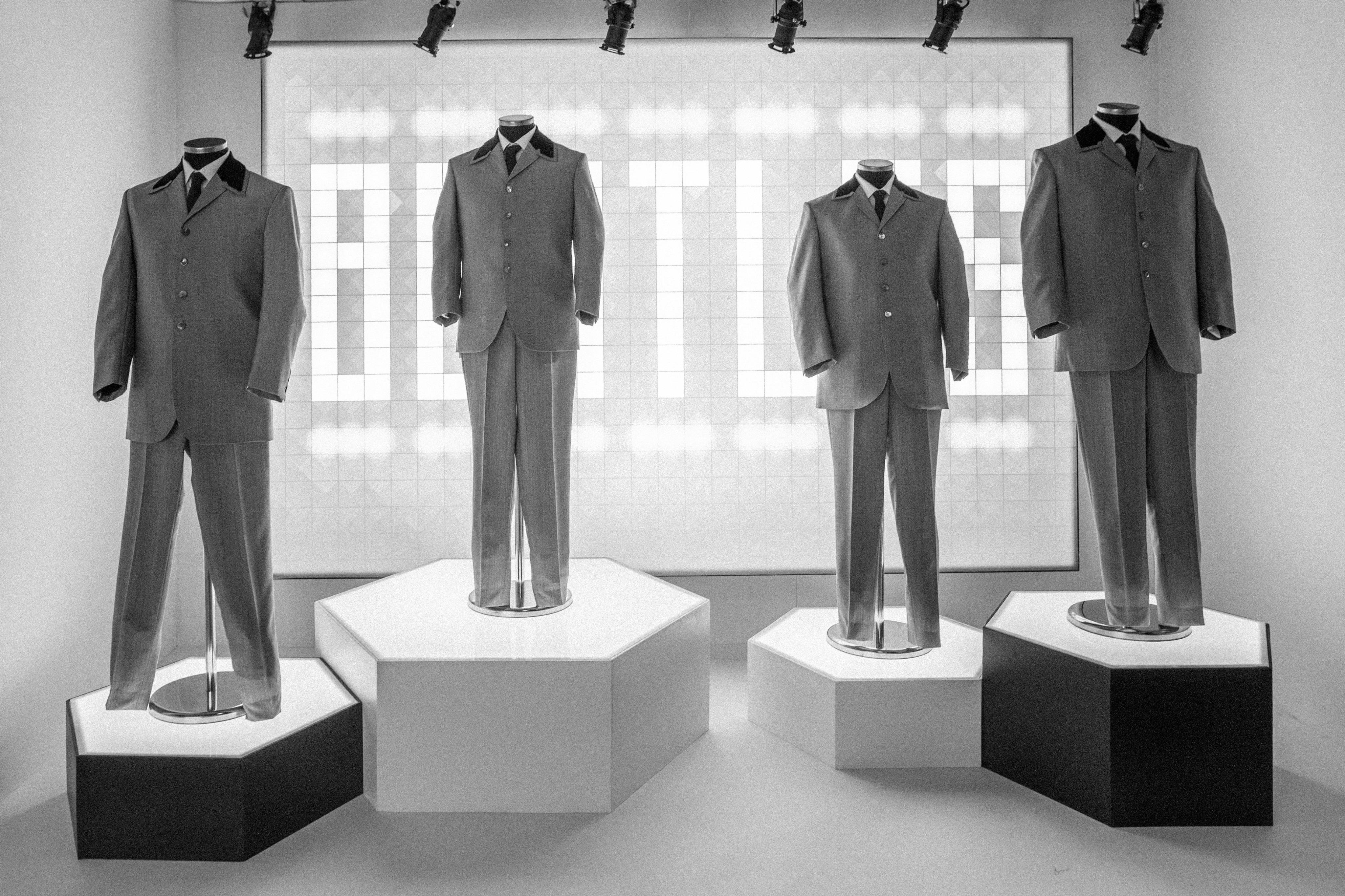 Fire mannequiner klædt i 1960'er-stil formelle dragter, der vises på piedestaler med "Beatles" oplyst i baggrunden.