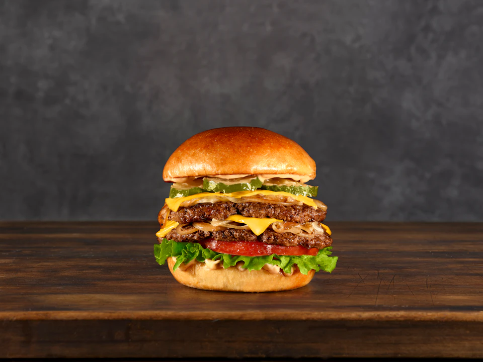 A double cheeseburger with lettuce, tomato, pickles, and cheese on a wooden surface against a dark background.