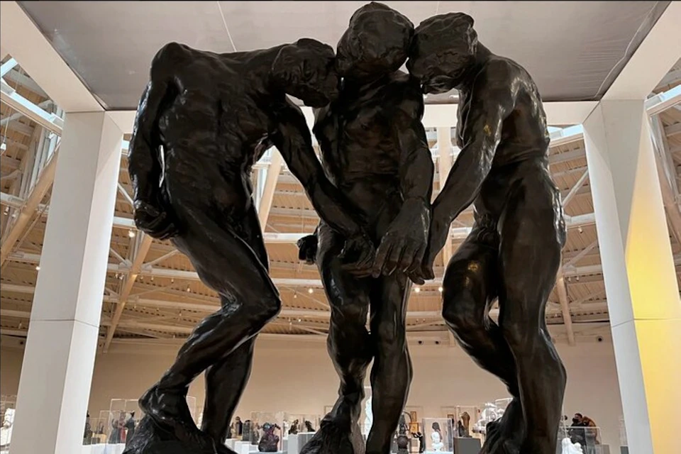 Three intertwined, dark bronze statues of human figures are displayed in a well-lit museum gallery.