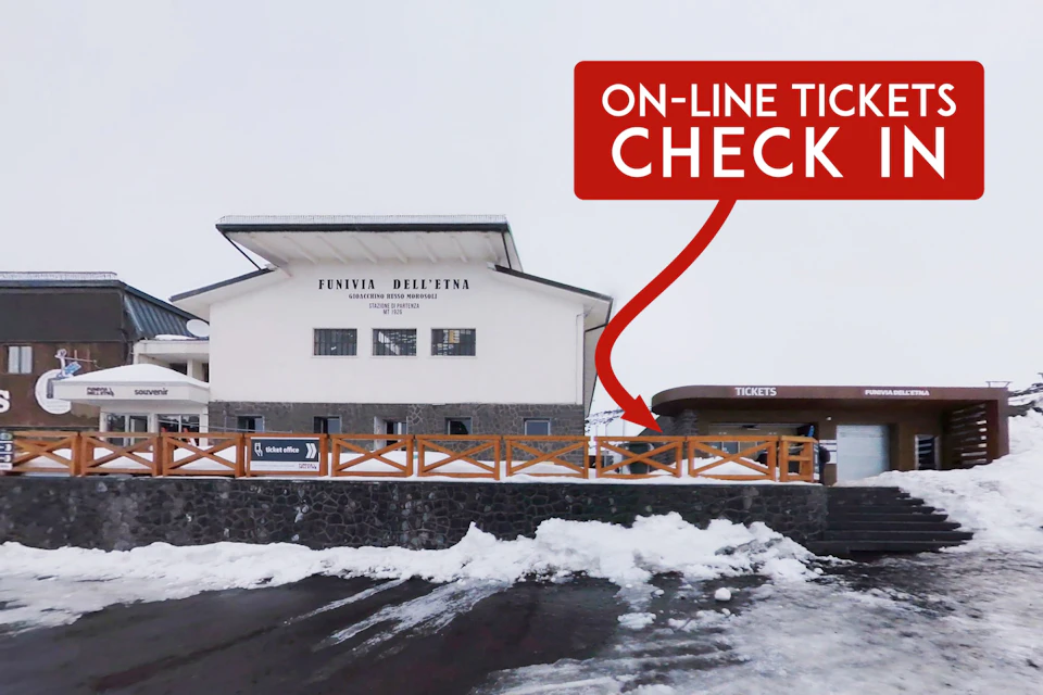 Snow-covered building labeled "Funivia dell'Etna" with a sign directing to an online ticket check area against a cloudy sky.