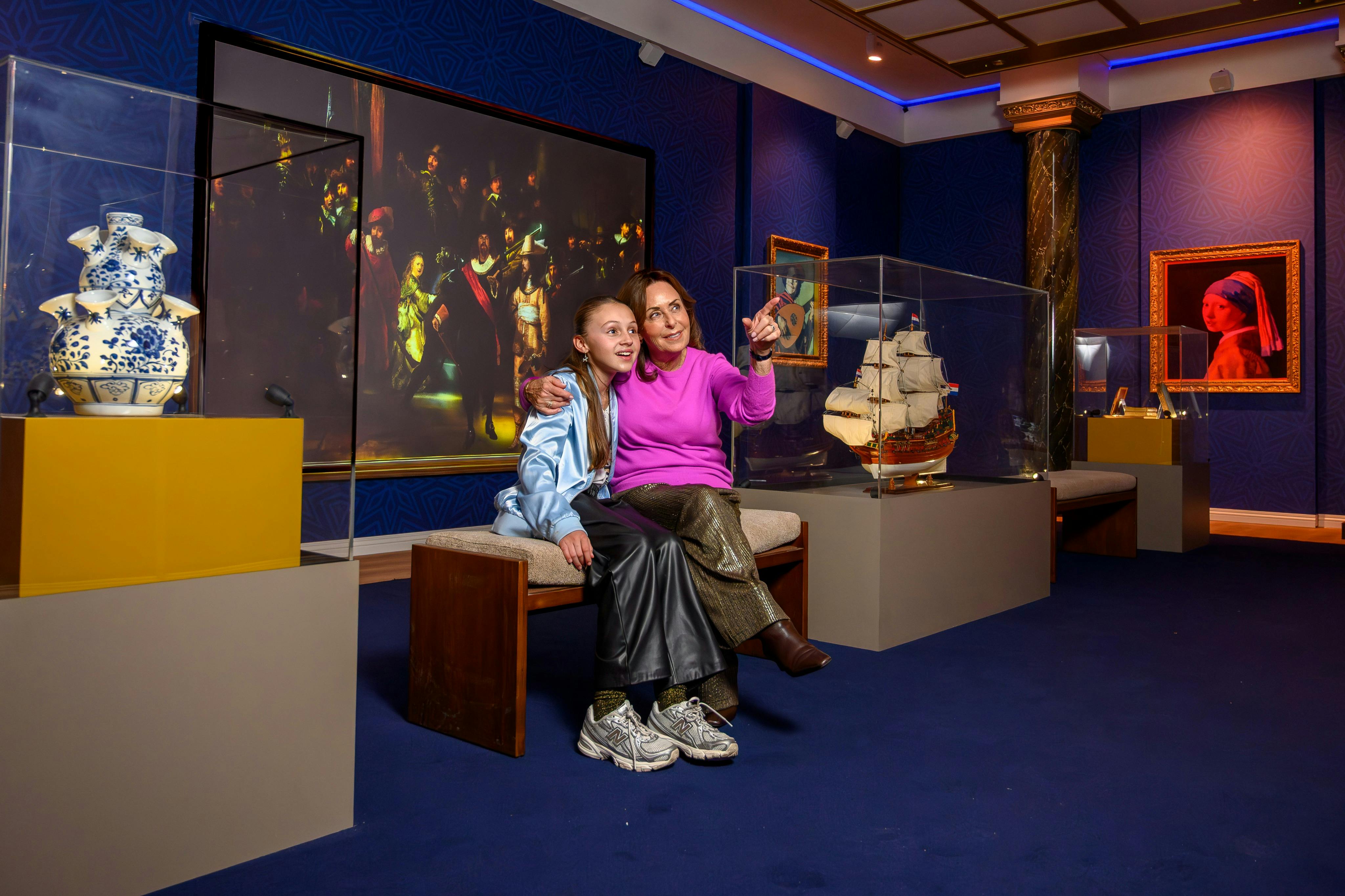 Två personer sitter på en bänk i ett museum, omgiven av montrar med konst och artefakter, inklusive en skeppsmodell och vas.