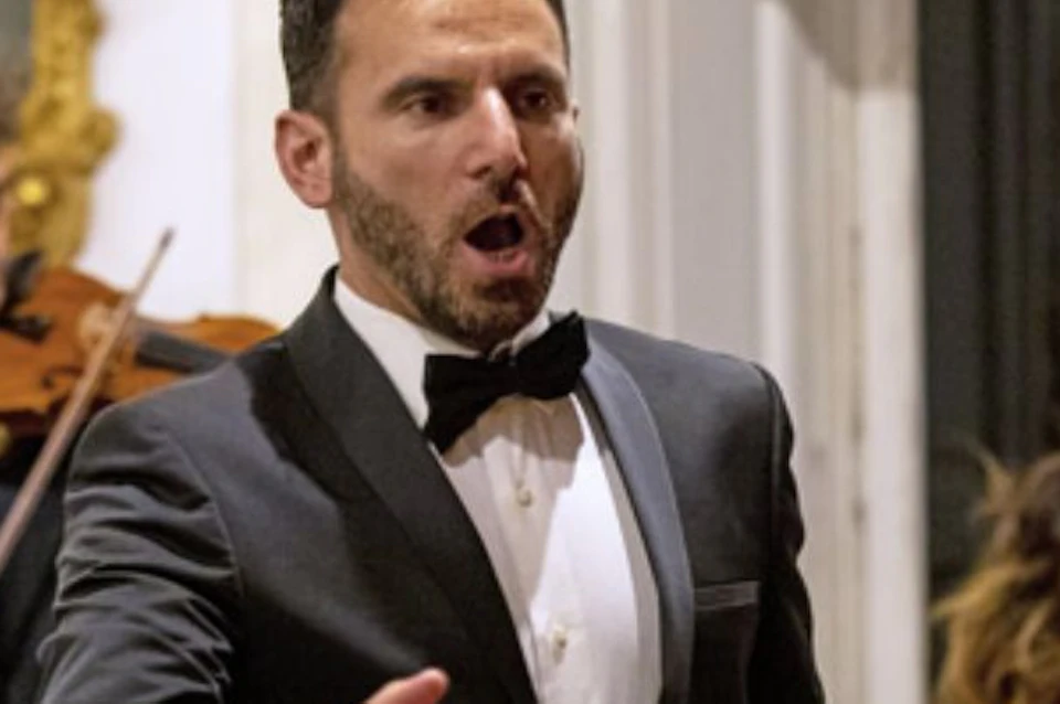 A man in a black tuxedo sings passionately, with a violinist and cellist performing in the background indoors.