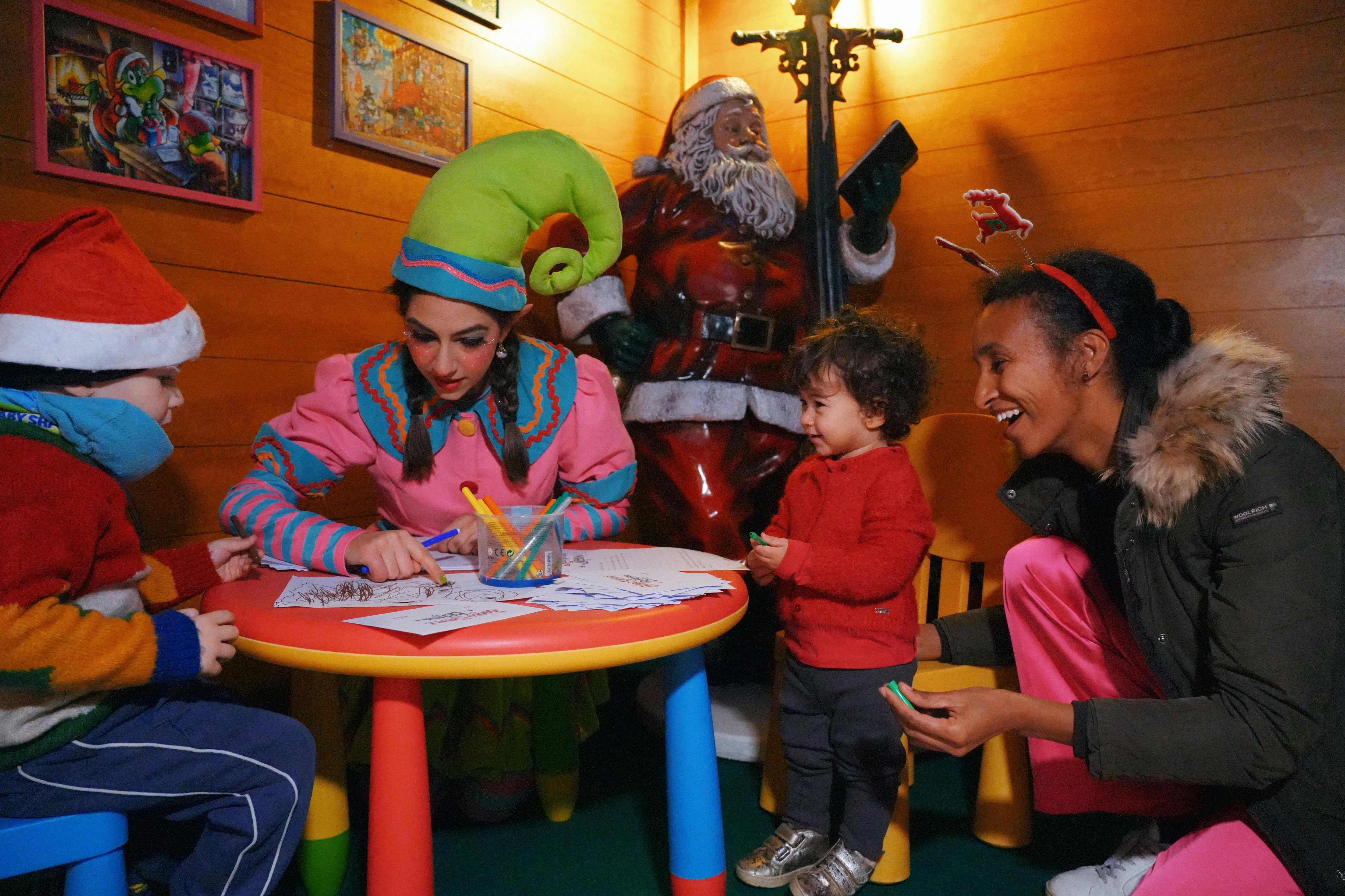 Osoba oblečená jako elf pomáhá dvěma dětem s barvením u stolu se sochou Santy v pozadí.