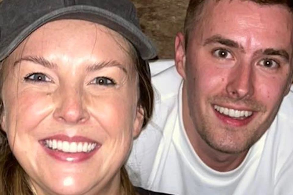 Two people smiling inside a narrow, rocky tunnel. One wears a cap and black shirt, the other a white shirt.