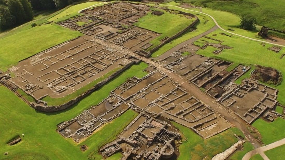 Aerial view of ancient stone ruins consisting of rectangular and grid-like structures, surrounded by green grass and paths.