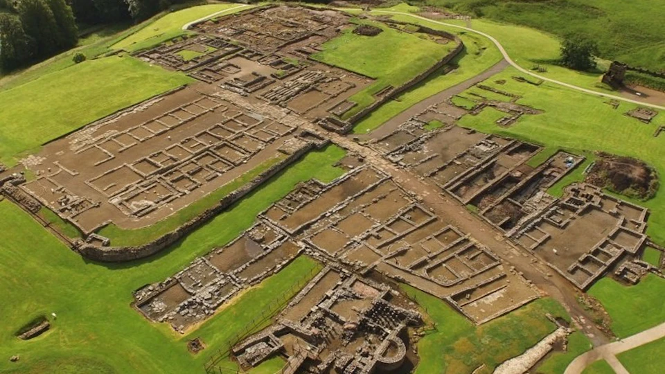 Aerial view of ancient stone ruins consisting of rectangular and grid-like structures, surrounded by green grass and paths.