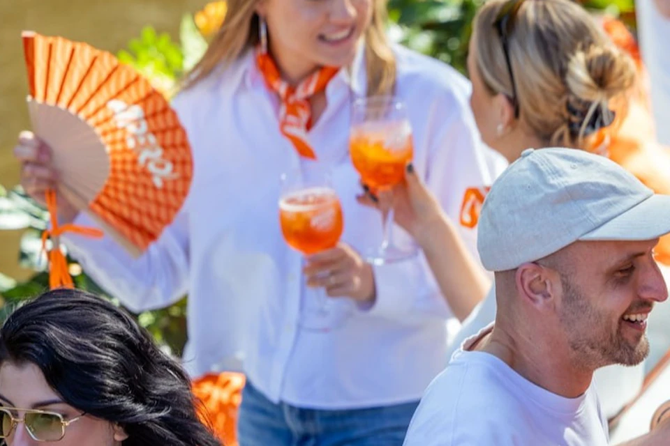 A group of people socializing outdoors, holding orange drinks; one woman holds an orange fan, and flowers are in the background.