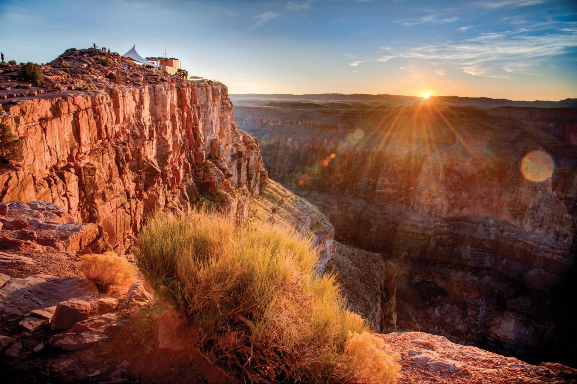 Sunrise over a canyon with rocky cliffs and sparse vegetation, featuring a building with a tent-like structure on the edge.