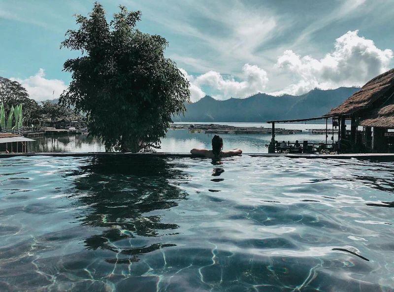 A person relaxes in an infinity pool overlooking a serene lake and mountains under a partly cloudy sky.