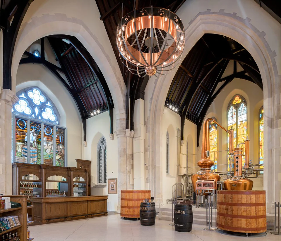 An interior of a converted church features large arched windows, a chandelier, distillery equipment, and wooden barrels.