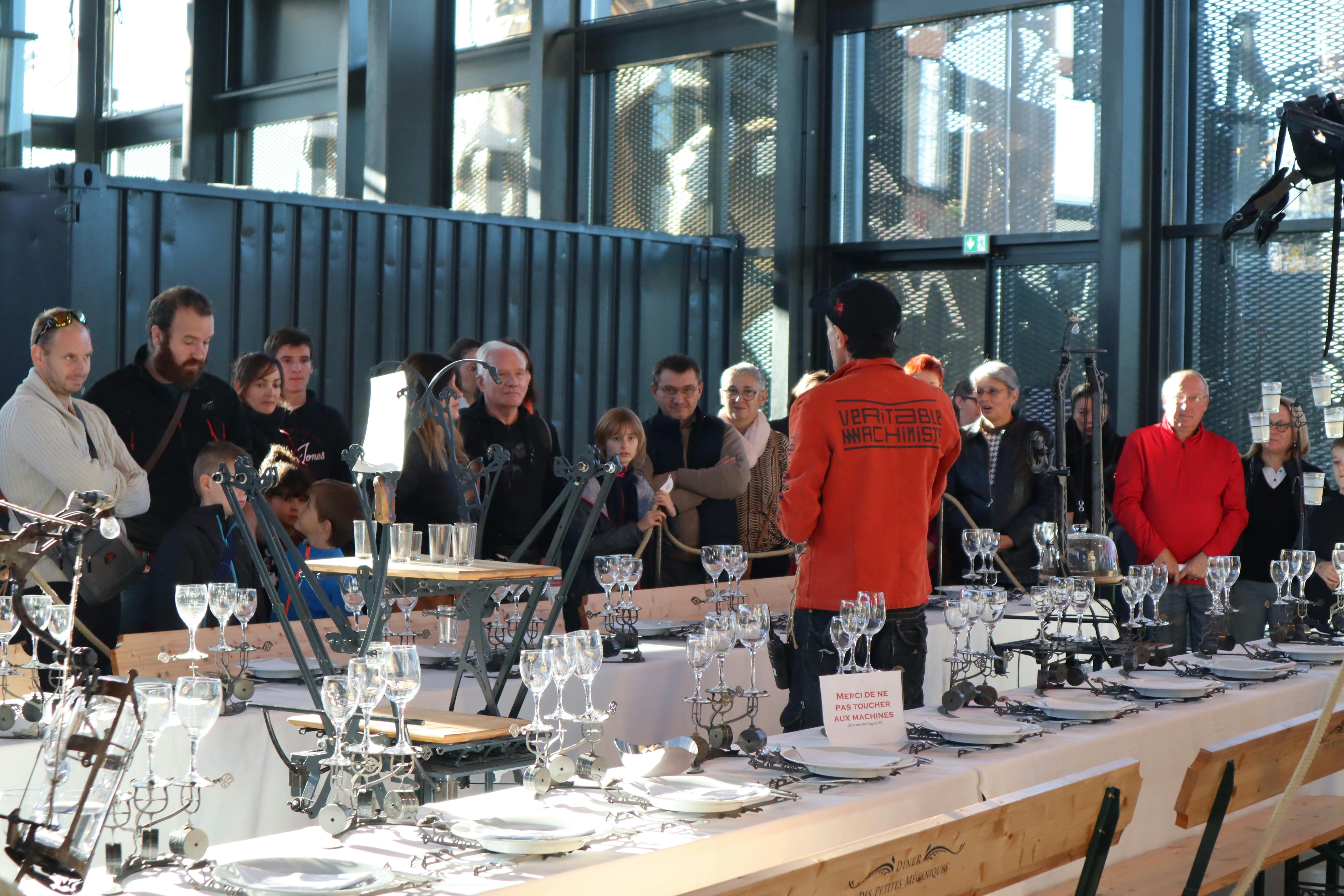 人们聚集在室内，观看一场演示，演示中摆放着错综复杂的桌子，上面摆放着机械装置和玻璃器皿。