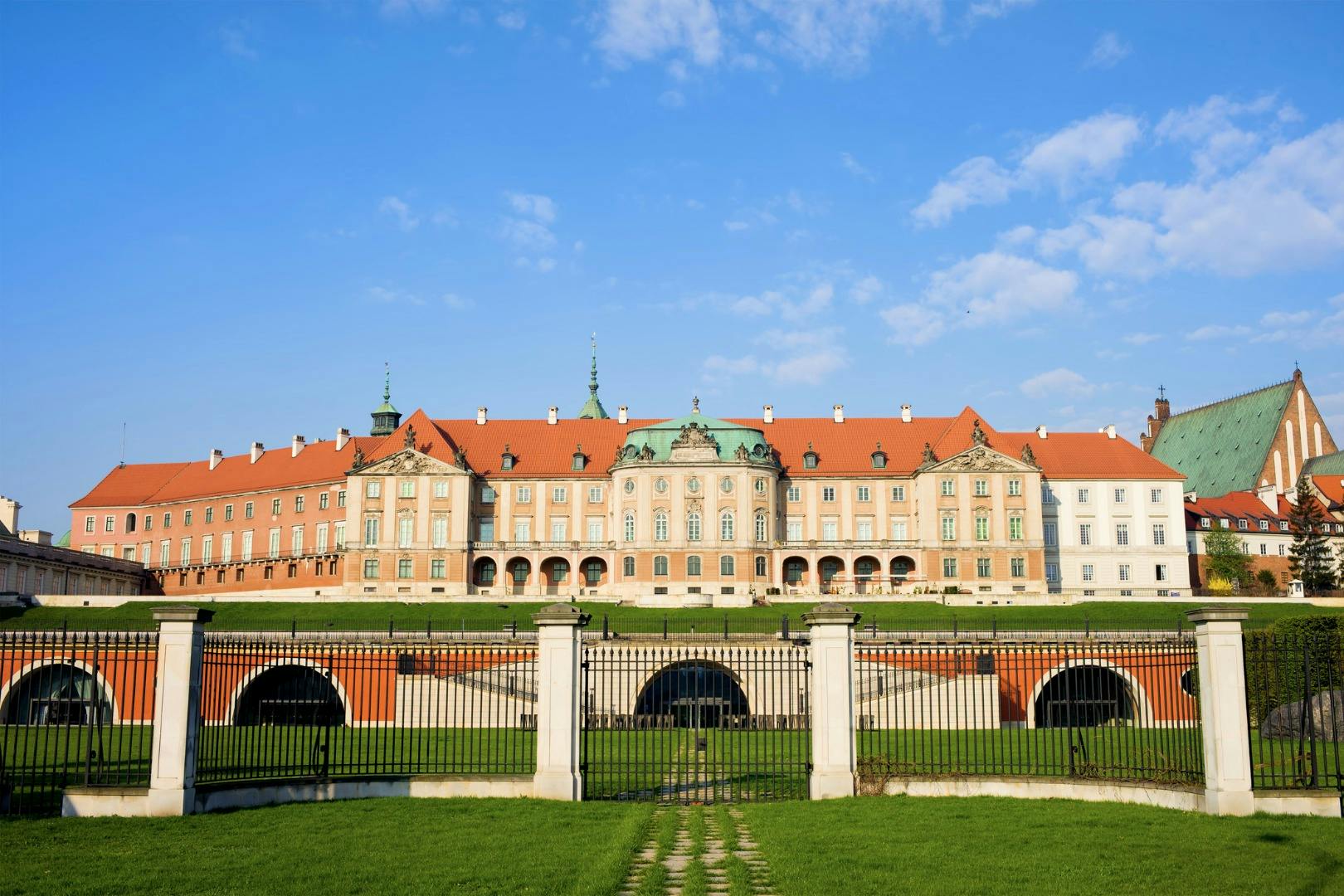 Ein großes historisches Gebäude mit roten Dächern und kunstvoller Architektur, hinter einem Rasen und einem dekorativen Zaun unter einem blauen Himmel.