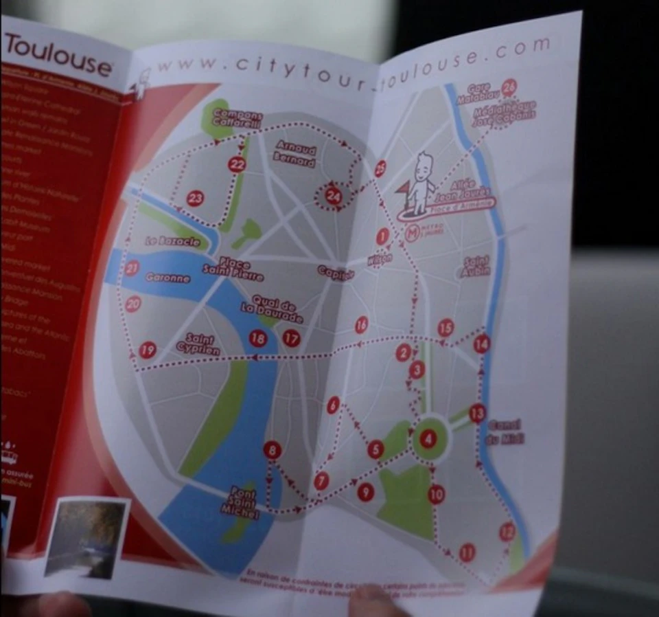 A person holding a city tour map of Toulouse, showing numbered landmarks and routes in red, on a background of blue and green areas.
