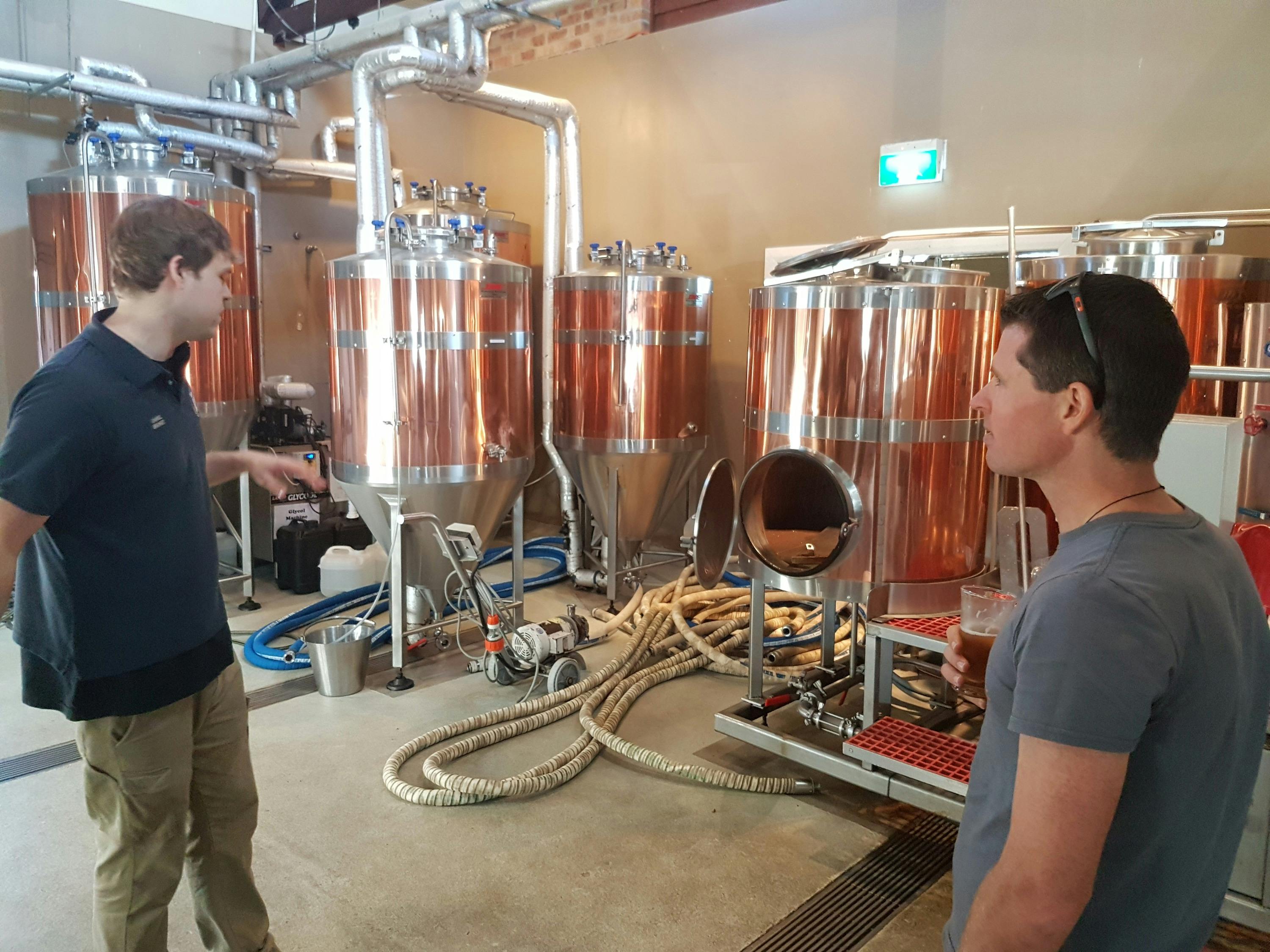 Two people in a brewery standing near large copper brewing tanks and equipment, surrounded by hoses and pipes.