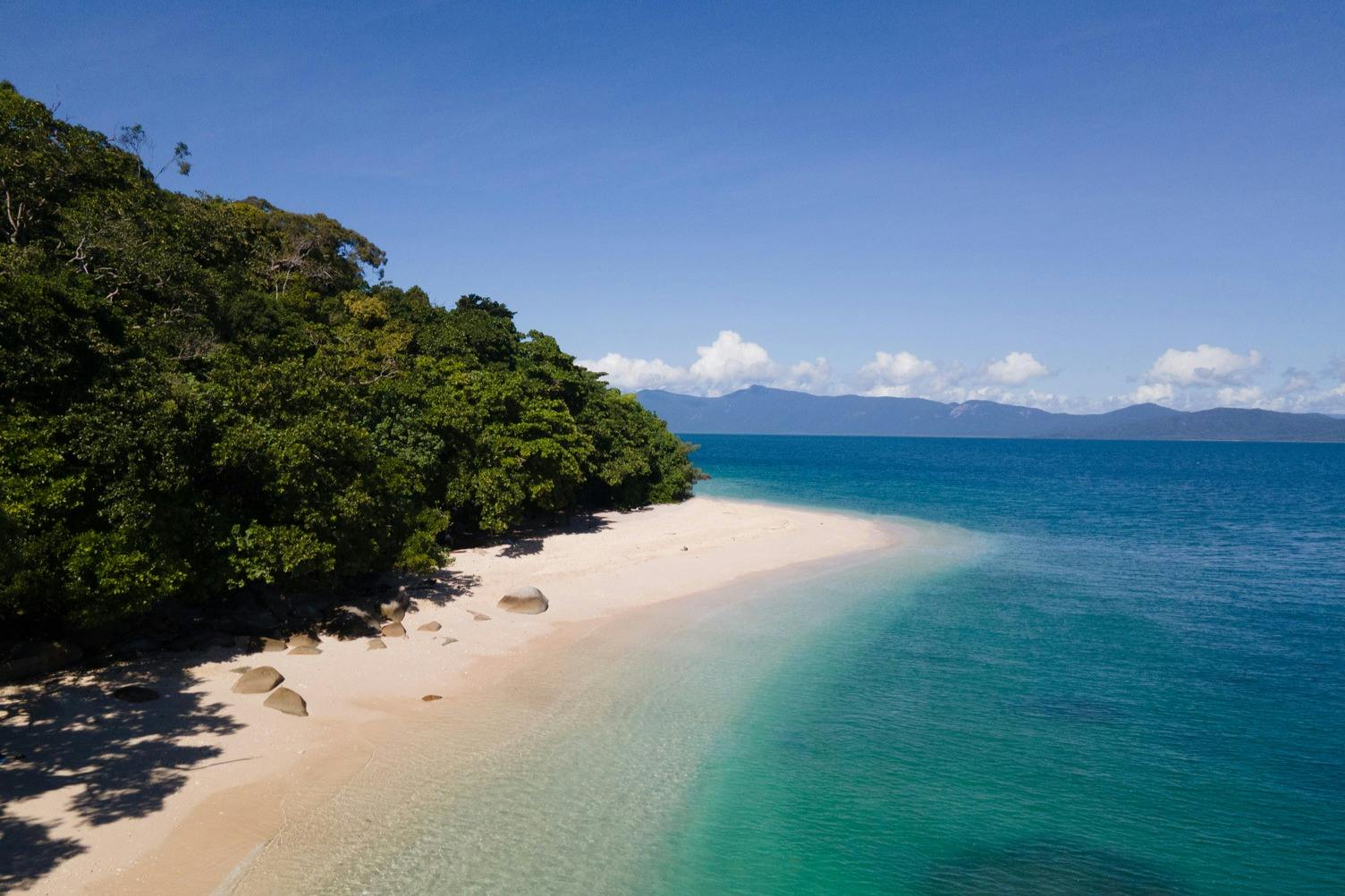 하얀 모래사장과 맑은 청록색 바다, 커다란 바위, 푸른 하늘 아래 푸른 나무가 우거진 한적한 해변.